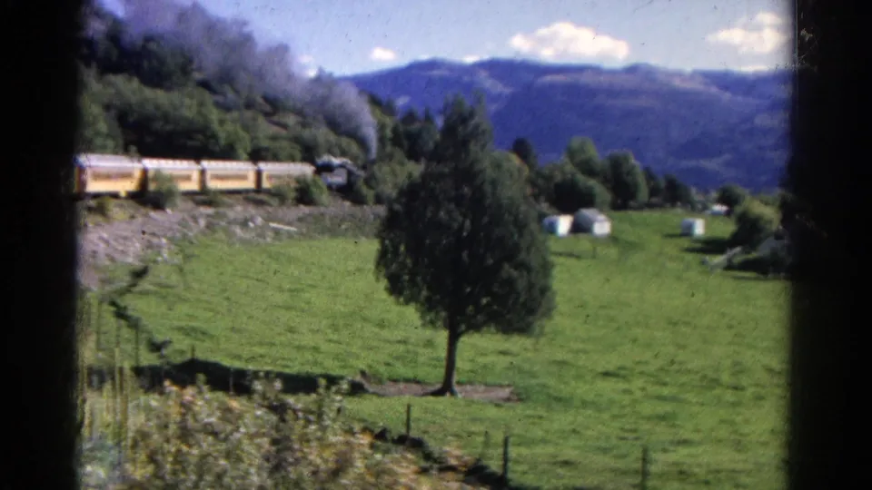 1967 Colorado 8mm frame with the train partly obscured by trees as it moves through a green valley below mountains.
