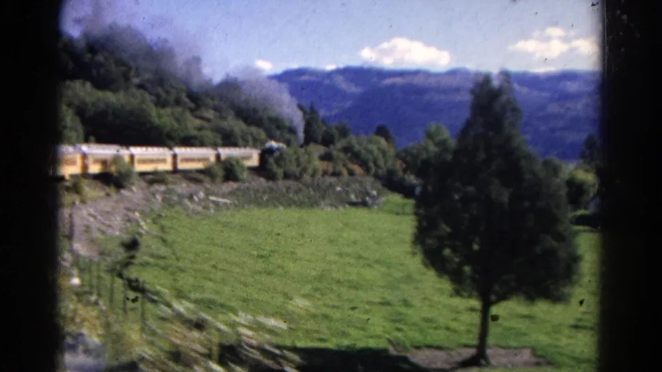 1967 Colorado home-movie frame showing a steam train on the hillside beyond a green field, with smoke and distant blue mountains.