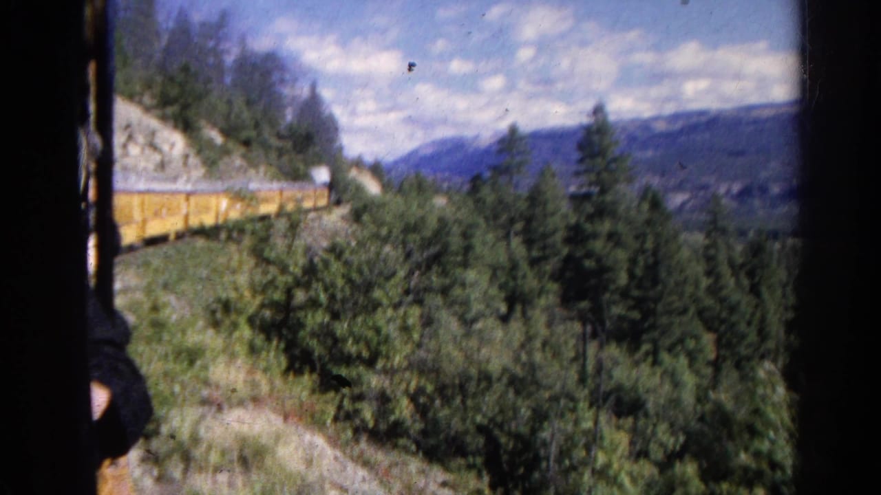 Yellow train curving past forest and rocky hillside with distant mountains.