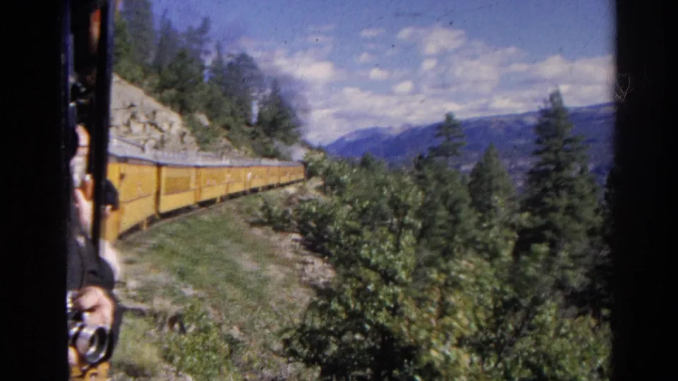 1967 Colorado train footage with yellow cars, mountain forest, and a passenger hand holding a small camera near the window frame.
