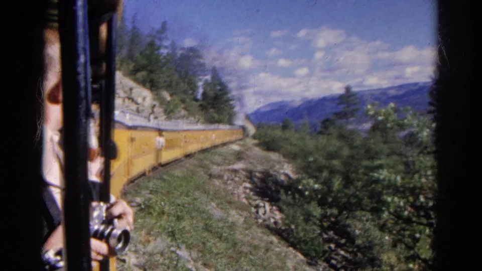 Passenger-view 8mm frame of a yellow train moving along a Colorado mountainside, with camera, hand, trees, rocks, and distant hills visible.