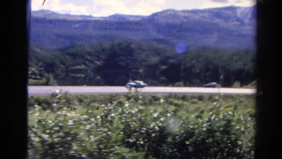 1967 Colorado 8mm frame showing a paved road with passing cars, grassy foreground, shrubs, and forested mountains in the distance.