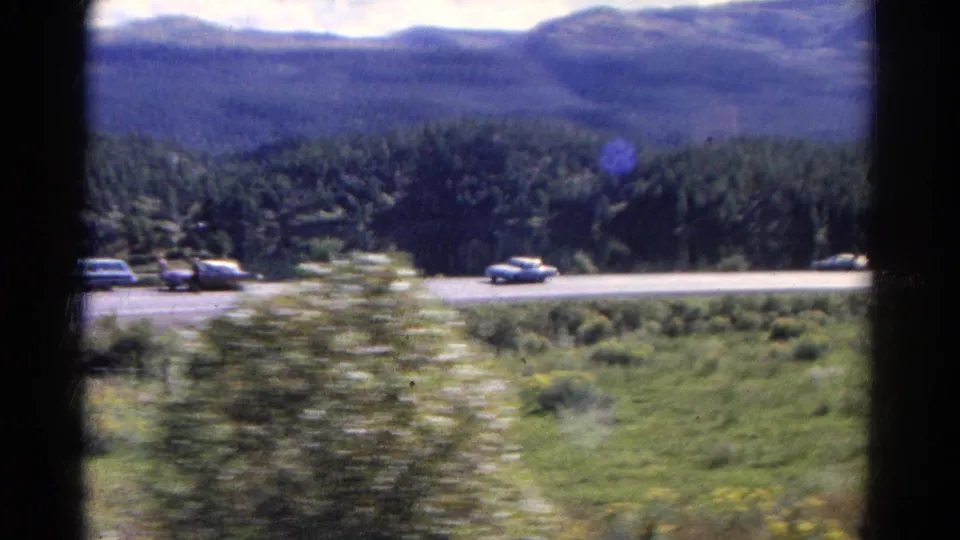 1967 Colorado home-movie frame of several cars on a mountain road, with shrubs, grass, dense forest, and distant blue hills.