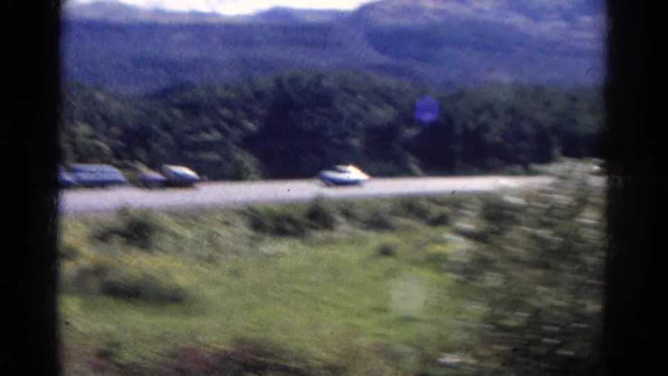 1967 Colorado 8mm frame from a moving vehicle, showing a road with several cars, grassy foreground, trees, and distant hills.