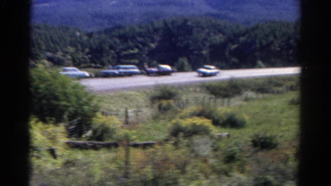 Cars on a roadside with trees, grass, fence posts, and hills behind.