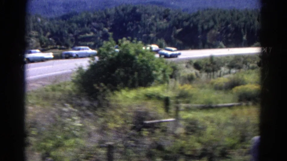 Moving 8mm view of a Colorado road with mid-century cars, roadside brush, fence lines, trees, and hills beyond.