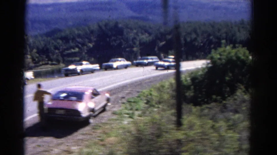 A roadside figure stands near a parked car in 1967 Colorado, with additional cars on the road and wooded hills behind.