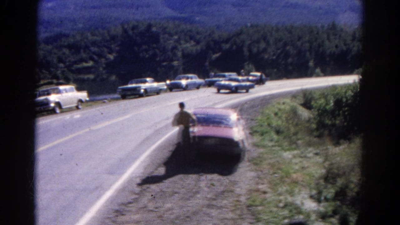 Curving mountain highway with period cars and a man near a parked car.