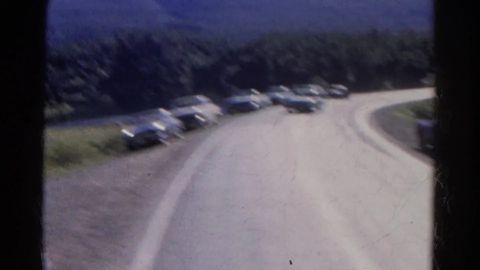 Soft 8mm train-window view of a Colorado mountain road with several cars along the curve and forested slopes beyond.