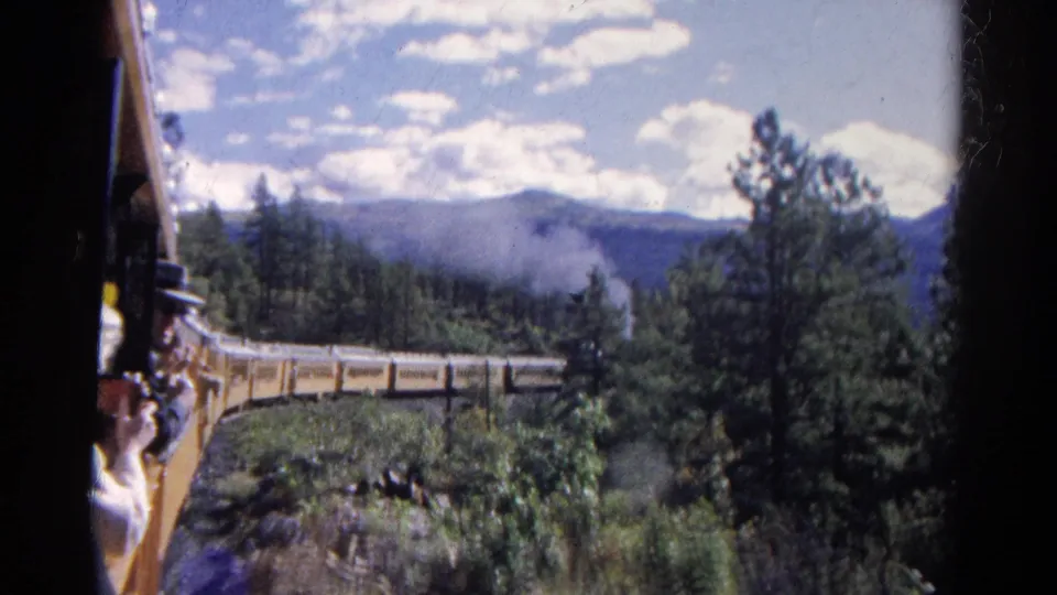 View from the train in Colorado with railcar edge, passengers, smoke, evergreen trees, and distant mountains under a bright sky.