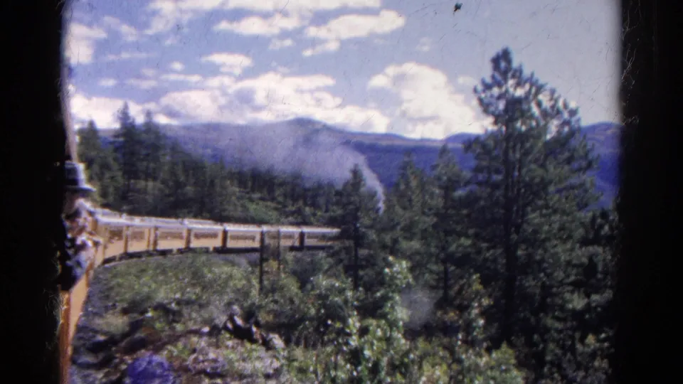 1967 Colorado 8mm frame showing a passenger at a train window with a train, bridge, pine trees, and distant mountains beyond.