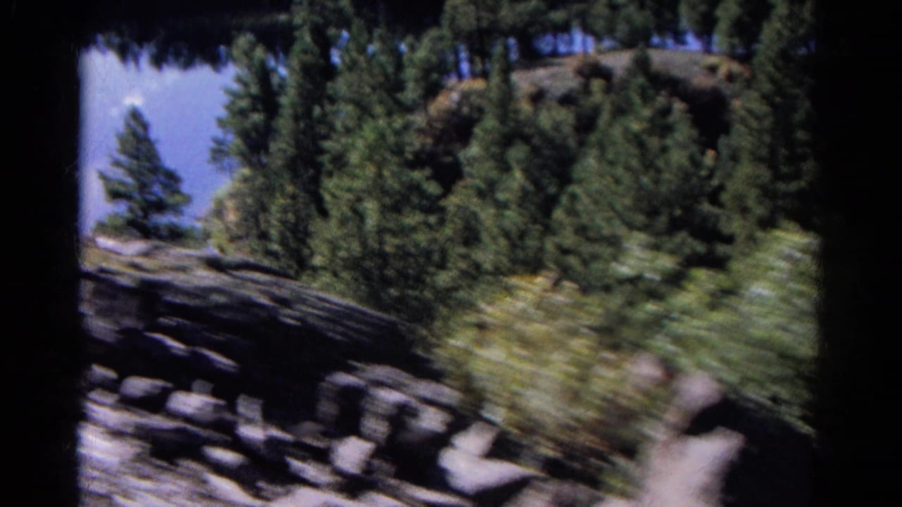 Moving 8mm view from a Colorado train, with rocks, pine trees, and hillside scenery blurred by speed and handheld motion.