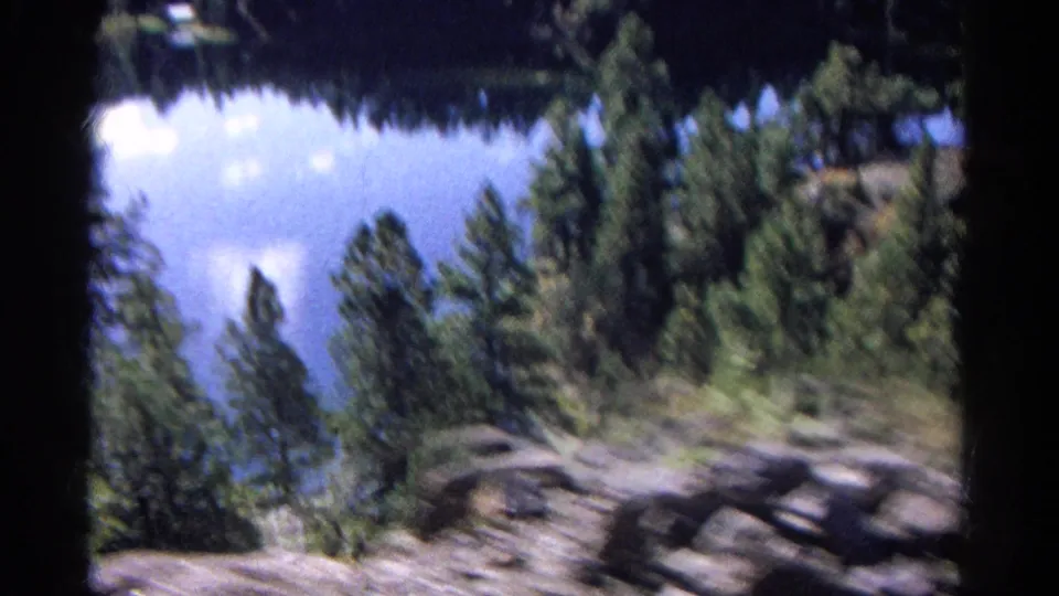1967 Colorado home-movie view of a lake through trees, with rocky terrain and evergreens streaking past the train.