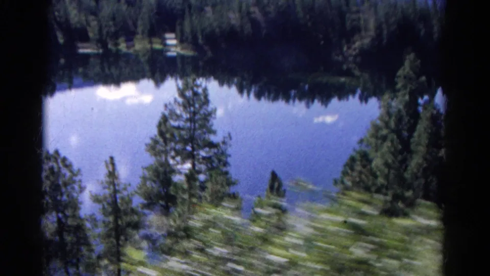 Colorado 8mm frame with a reflective lake, pine-covered shore, sky reflections, and blurred foreground vegetation from the moving train.