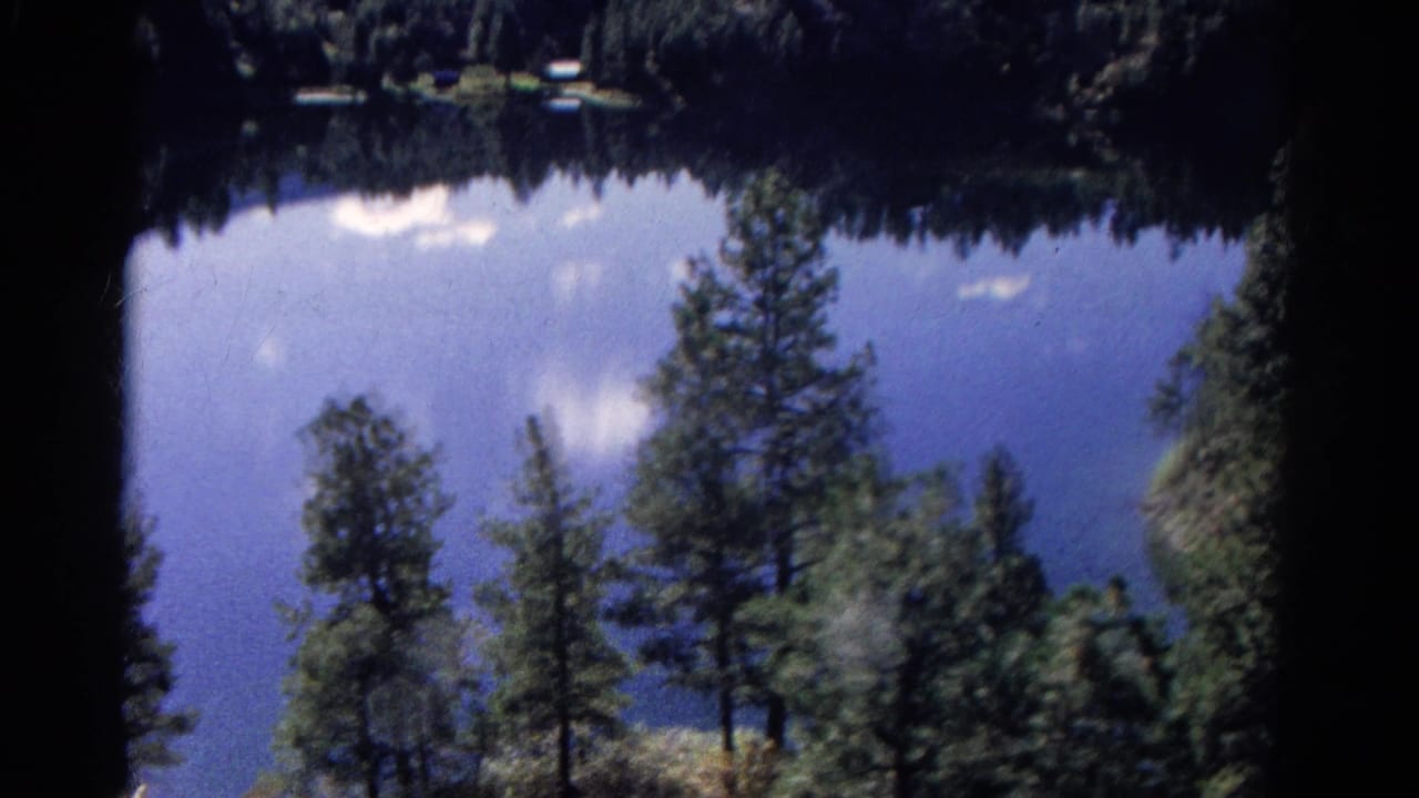 1967 Colorado 8mm home-movie view of pine trees beside a blue mountain lake with reflected sky and dark film borders.
