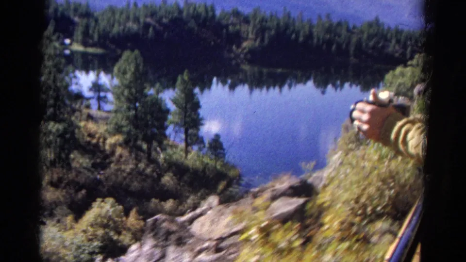 1967 Colorado 8mm lakeside overlook with pine forest, rocky slope, blue water, and a partial hand holding a camera at the right edge.