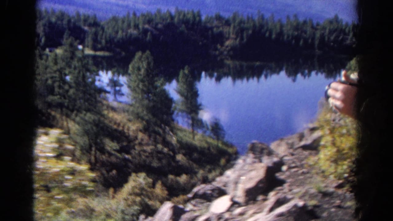1967 Colorado 8mm frame showing a blue mountain lake, evergreen shoreline, rocky slope, and a partial hand at the edge.