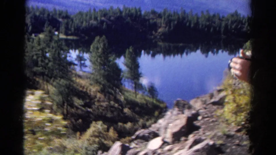 1967 Colorado 8mm frame showing a blue mountain lake, evergreen shoreline, rocky slope, and a partial hand at the edge.