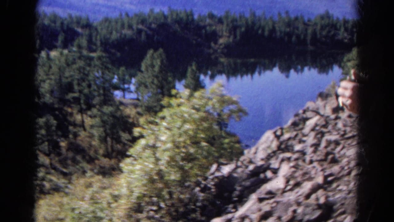 1967 Colorado 8mm frame of a calm lake below dense evergreens, with rocky terrain and foreground brush.