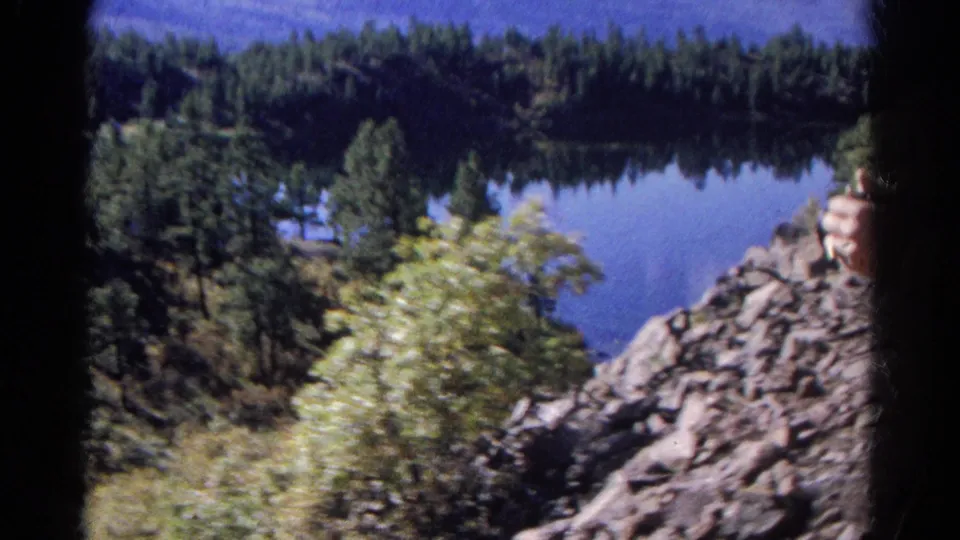 1967 Colorado 8mm frame of a calm lake below dense evergreens, with rocky terrain and foreground brush.