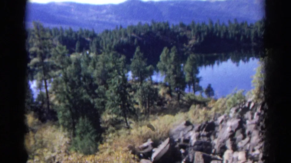 1967 Colorado home-movie frame showing pine trees, dry brush, rocks, and a blue lake below the hillside.