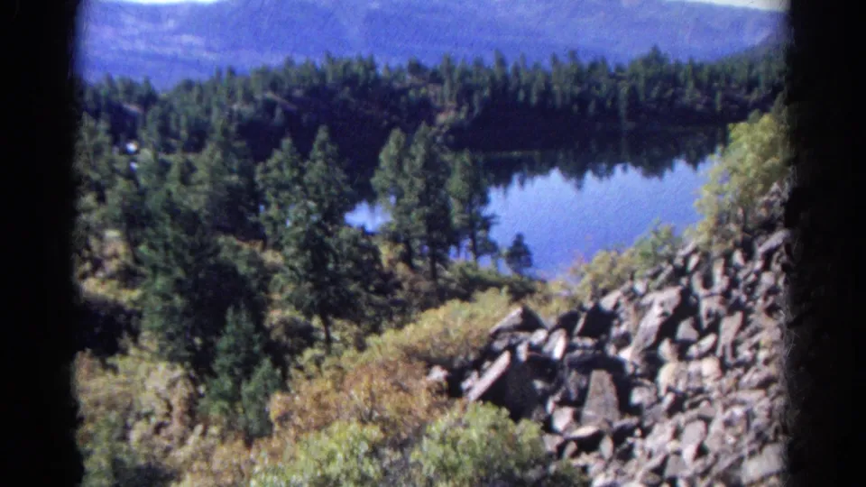 1967 Colorado 8mm frame with rocks, grasses, pines, and a calm lake set below a wooded ridge.
