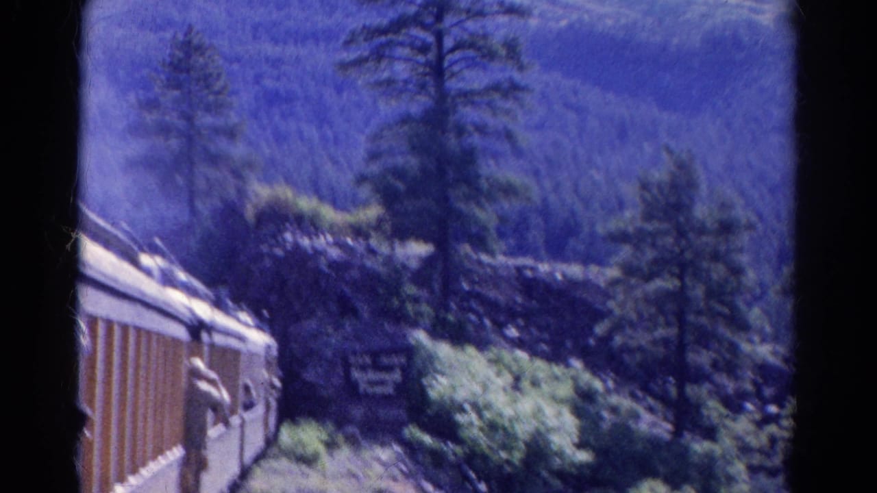 1967 Colorado home-movie frame showing a passenger train beside wooded mountain terrain and a San Juan National Forest sign.
