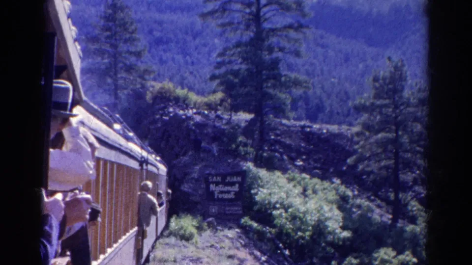 1967 Colorado 8mm frame with passengers visible along a train window line beside a San Juan National Forest sign and wooded slopes.