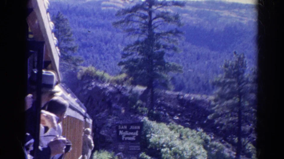 Final 1967 Colorado train-travel frame with riders at open windows, pine forest, mountain slopes, and a readable San Juan National Forest sign.