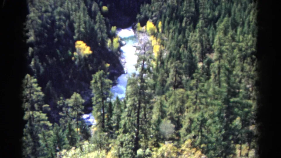 Elevated 8mm Colorado landscape frame showing a pale river bend, tall conifers, rocky banks, and autumn color in the trees.