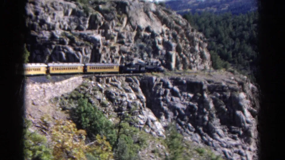 1967 Colorado 8mm frame of a yellow passenger train moving across a cliffside rail line above canyon vegetation.