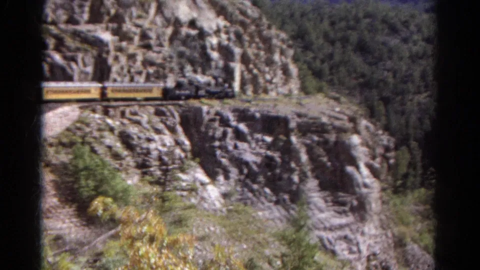 1967 Colorado mountain rail scene with yellow train cars crossing a rocky canyon ledge above trees and brush.