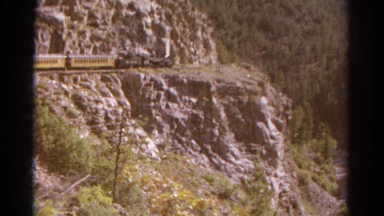 1967 Colorado 8mm frame showing a steam locomotive and yellow passenger cars on a narrow track beside a steep rock face.
