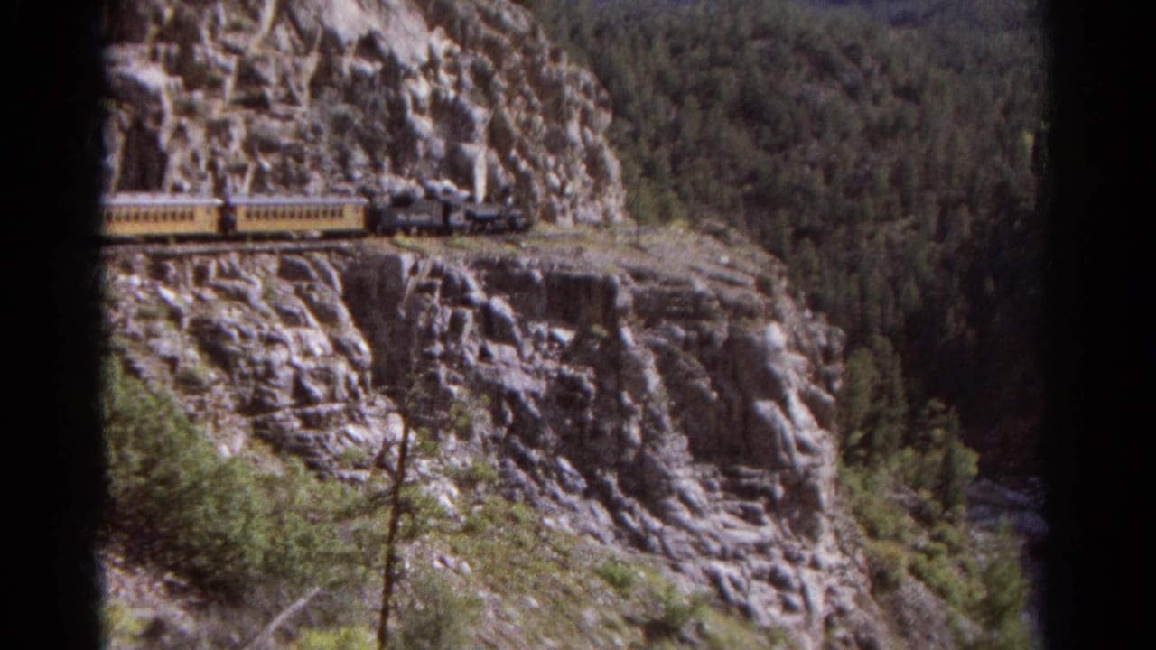 Steam train appears on a Colorado mountain ledge, with yellow passenger cars, jagged rock, and forested canyon beyond.