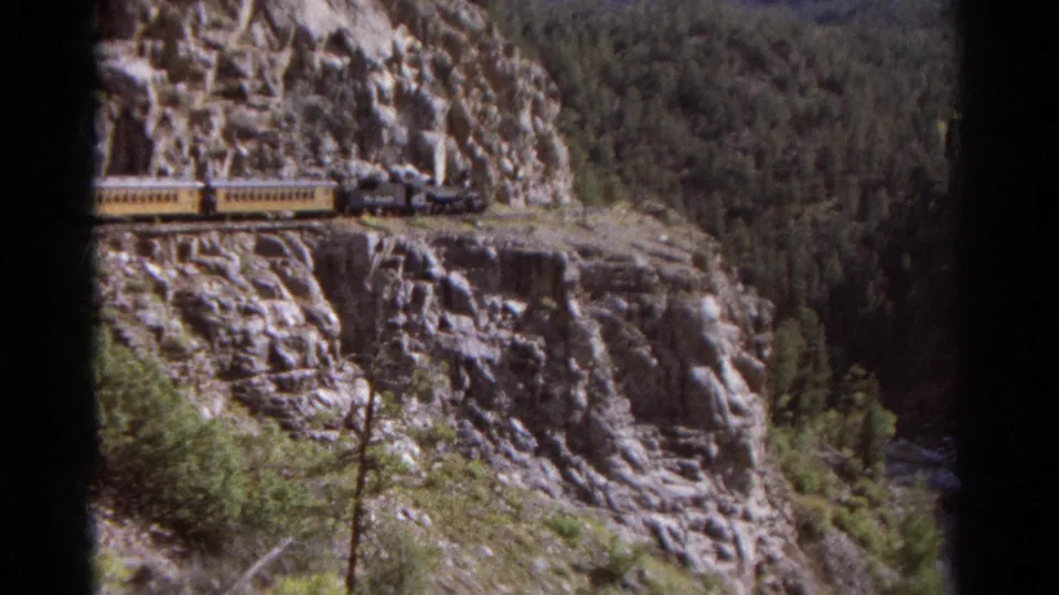 Steam train appears on a Colorado mountain ledge, with yellow passenger cars, jagged rock, and forested canyon beyond.