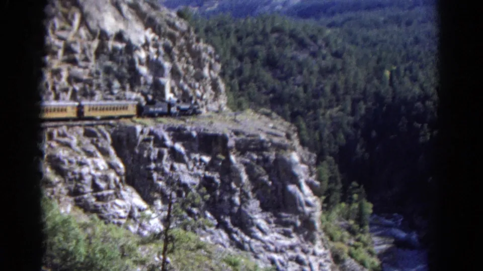 Colorado 8mm view of a steam train curving along a rocky mountainside, framed by trees, canyon shadow, and film edge darkness.