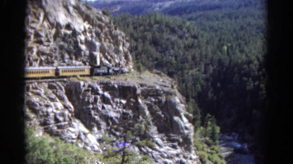 Late-1960s Colorado home-movie frame of a steam train on a cliffside curve, with yellow cars, rugged rock, and evergreen forest.