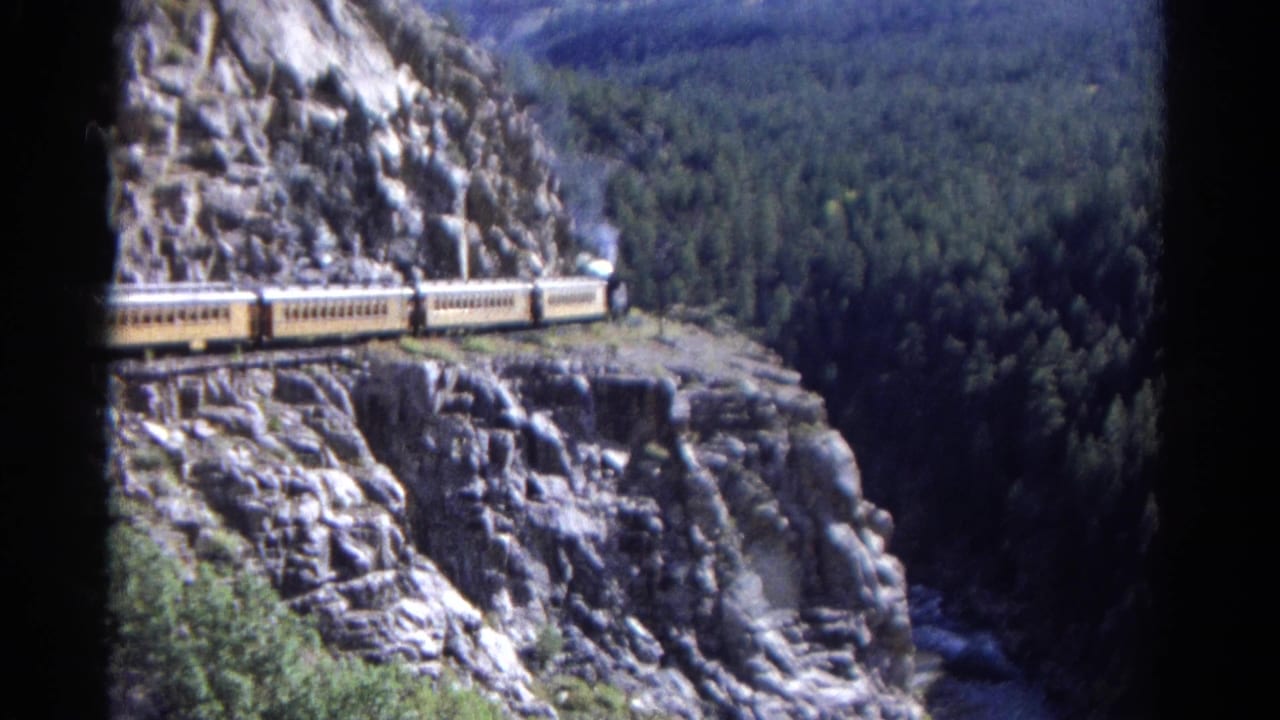 Steam train with yellow passenger cars crossing a rocky mountain ledge above forest.