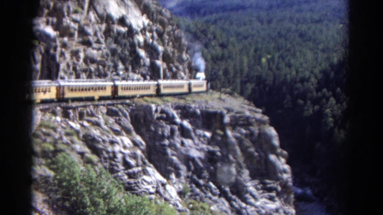 1967 Colorado home-movie frame showing yellow passenger cars on a ledge with steam smoke and dense forest in the background.