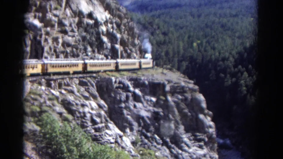 1967 Colorado home-movie frame showing yellow passenger cars on a ledge with steam smoke and dense forest in the background.