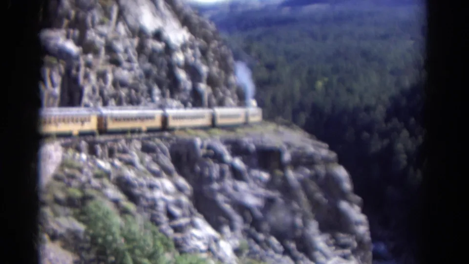 Soft-focused 1967 Colorado 8mm frame of a yellow passenger train crossing a rocky ledge above a forested drop.