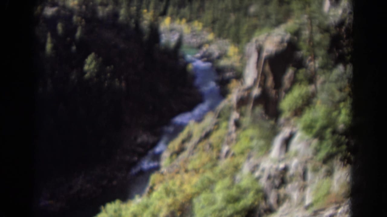 1967 Colorado 8mm frame showing a narrow stream below rocky canyon slopes with trees and sunlit vegetation.