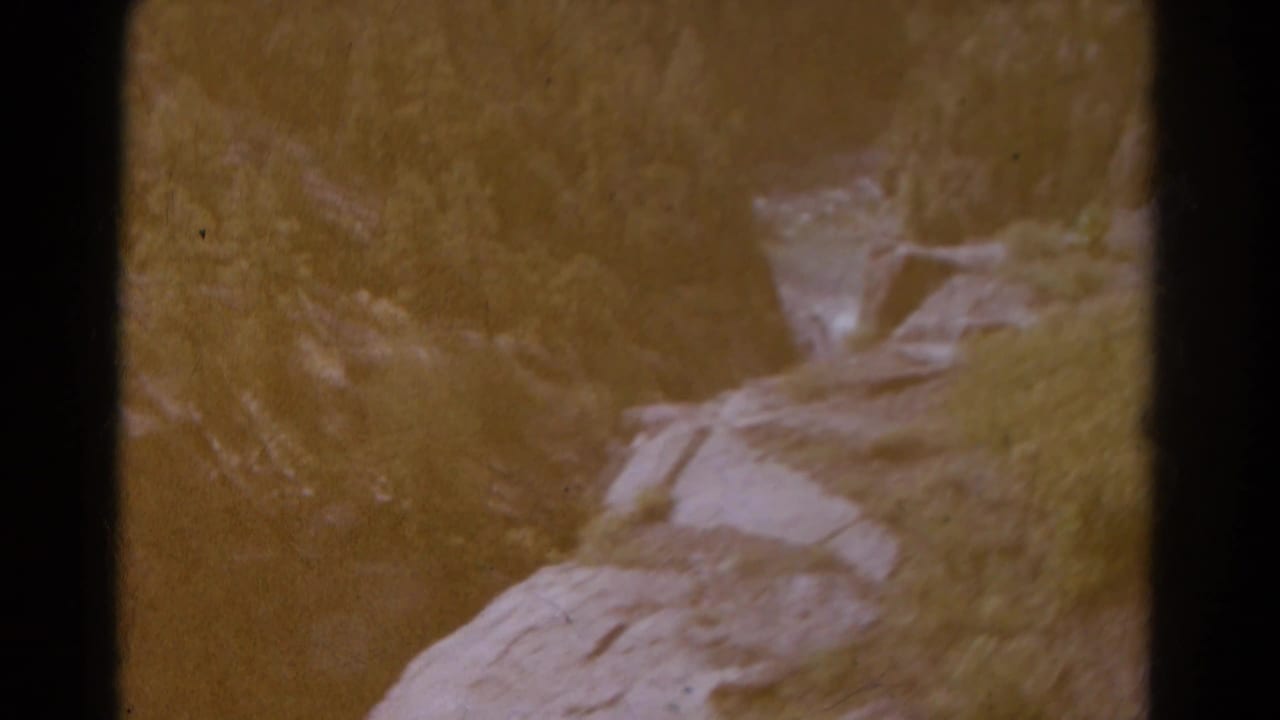 Soft 8mm view of pale rocks and grass along a shadowed mountain canyon edge.