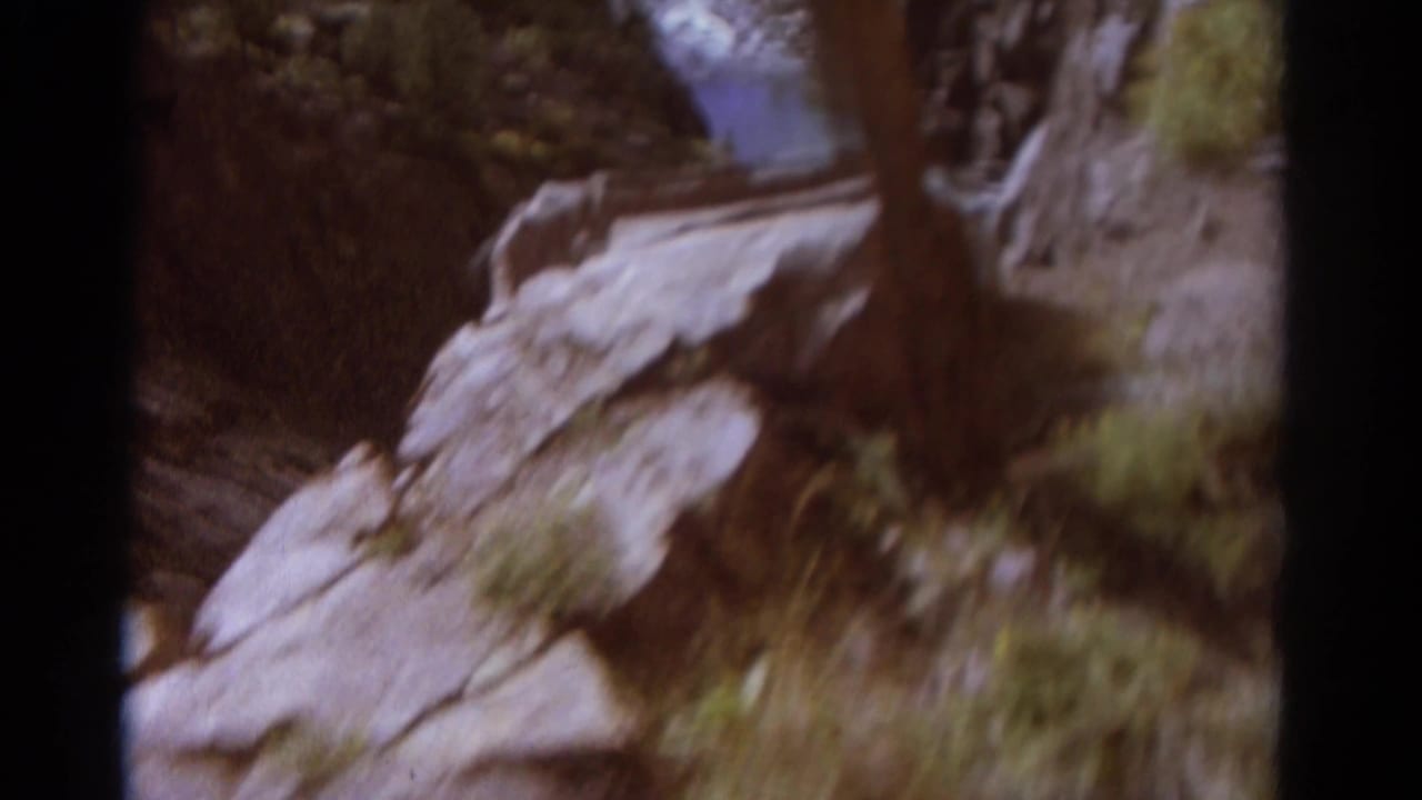 1967 Colorado home-movie frame looking down a rocky canyon slope toward shrubs, grass, and a thin stream in the ravine.