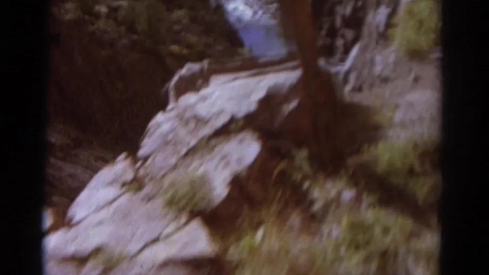 1967 Colorado home-movie frame looking down a rocky canyon slope toward shrubs, grass, and a thin stream in the ravine.