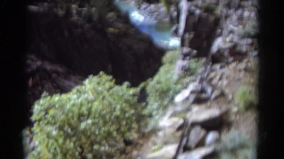 1967 Colorado 8mm frame showing a rocky canyon bottom, green shrubs, steep walls, and a narrow stream in soft home-movie focus.