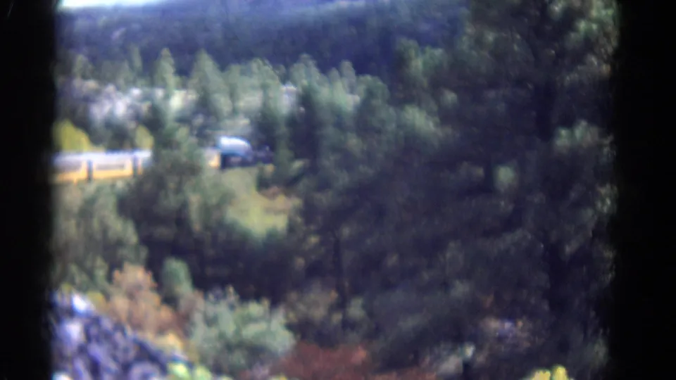 A yellow train passes behind trees above a rocky canyon and river area in vintage Colorado home-movie footage.