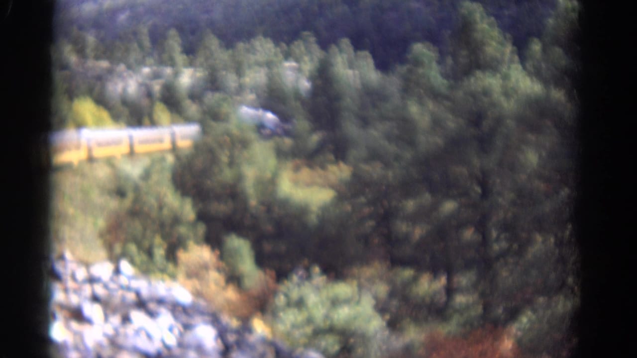 1967 Colorado 8mm frame showing a yellow passenger train crossing a wooded mountain slope above rocks and dense trees.