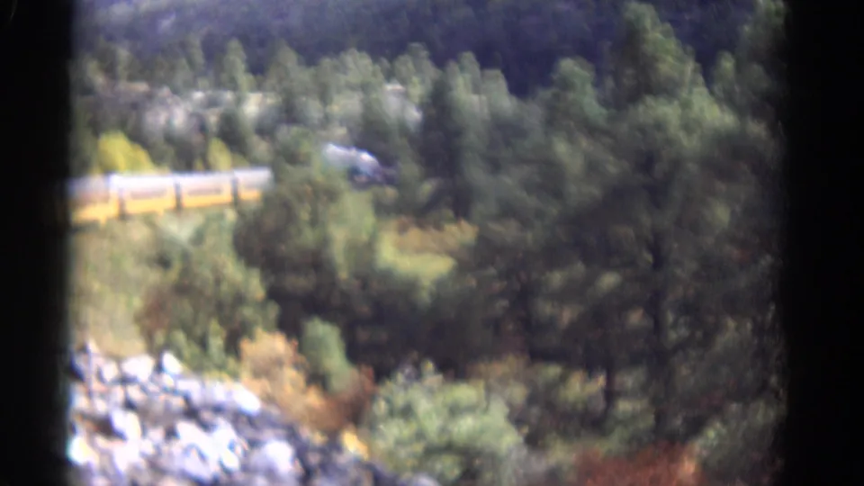 1967 Colorado 8mm frame showing a yellow passenger train crossing a wooded mountain slope above rocks and dense trees.