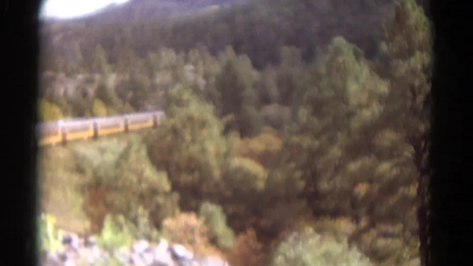 A 1967 Colorado home-movie view shows yellow train cars partly screened by trees above a rocky foreground.